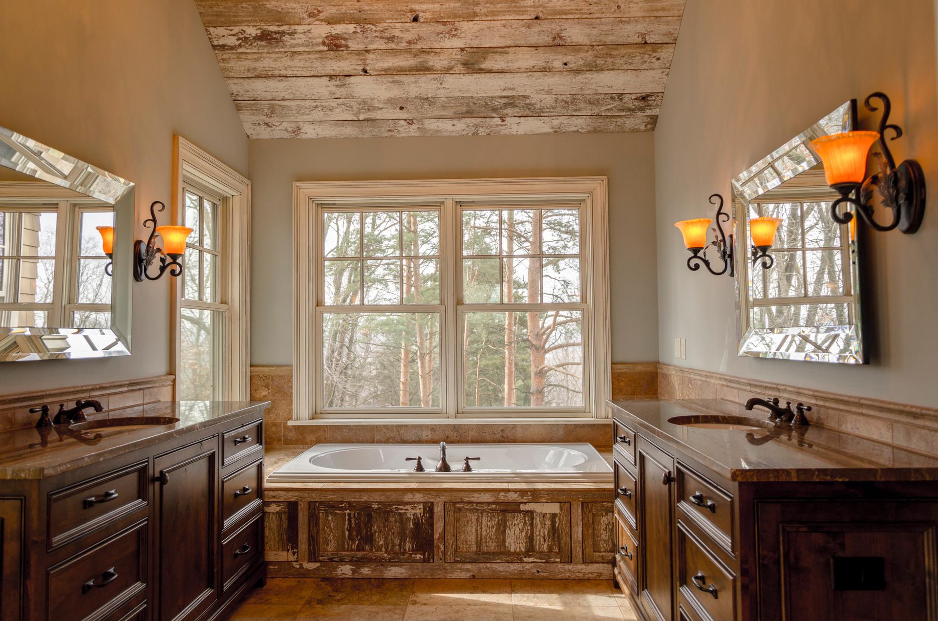 This bathroom features two vanities, a bathtub, warm lighting and three windows that overlook the outdoors.